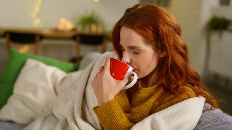 Young woman enjoying a cup of hot coffee as she snuggles under a blanket for warmth on a cold winter day | Kid Vs. Cold: The True Story of the Best Blanket Ever