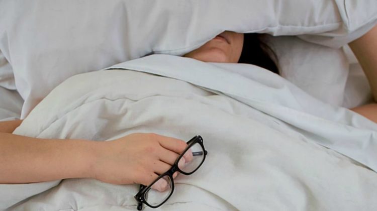 Woman sleeping in bed holding her glasses | Gadgets To Help You Fall Asleep Quicker | Featured